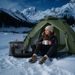 Person sitting in a green tent in a snowy mountain landscape with a portable heater and cup.