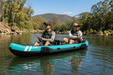 Two people fishing in a teal inflatable kayak on a calm river with trees and mountains in the background.
