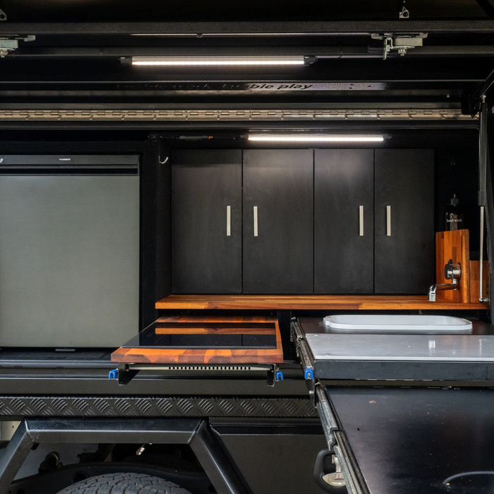 Storage compartment with black cabinets, wooden shelves, and a fridge/freezer inside a vehicle.