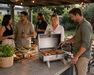 Group of people cooking on a BBQ outdoors with food and drinks.