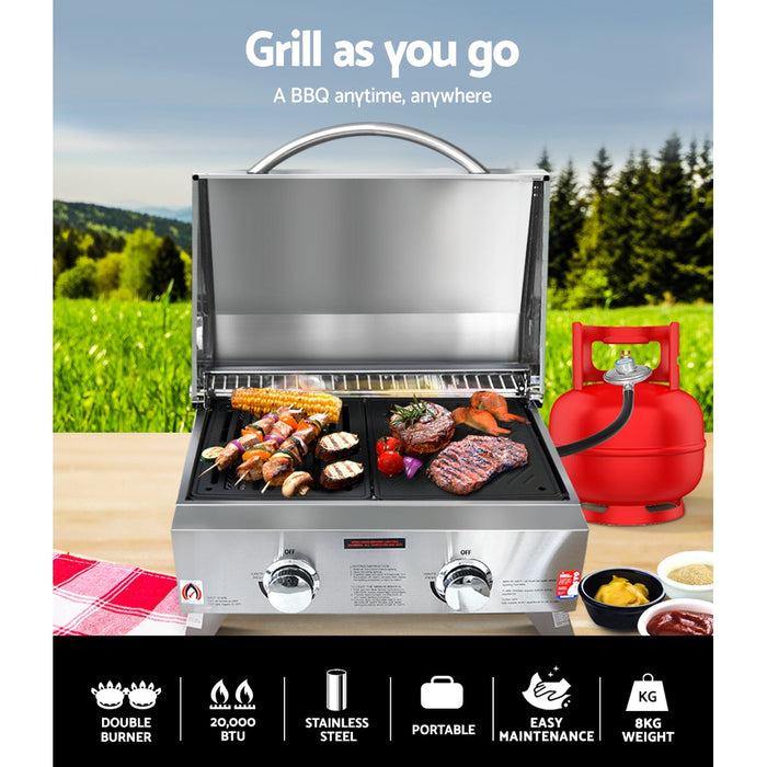 Portable gas grill with food on a table outdoors, accompanied by a propane tank.